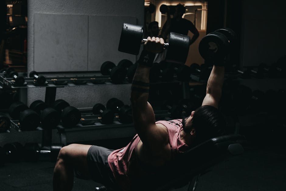 Muscular man in sportswear performing dumbbell bench press in a dimly lit gym setting.